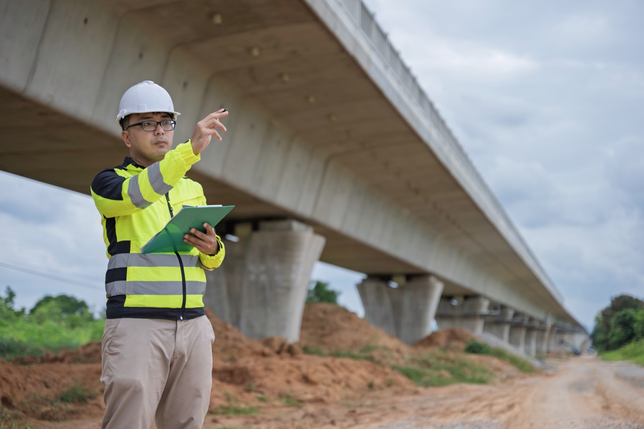 Construction engineer wearing safety gear and holding a tablet inspects bridge development site. The scene highlights infrastructure progress, civil engineering, and technology use in construction.